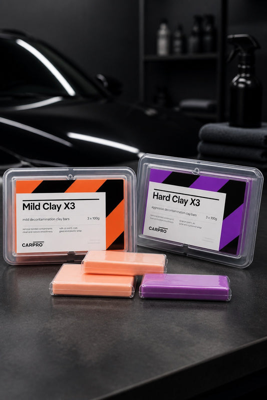 Envases de CarPro Mild Clay X3 y Hard Clay X3 con dos barras de arcilla sobre una superficie oscura y un coche al fondo.